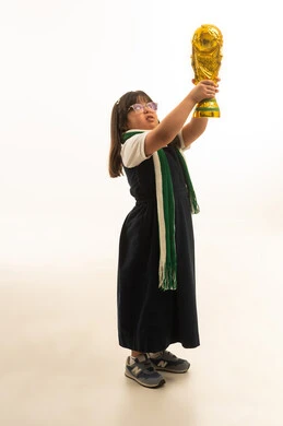 Raising the golden cup high after achieving victory, a small Arab Gulf Saudi girl with special needs wearing a gray school uniform and a green keffiyeh, looking up, full-length body, portrait with a white background.