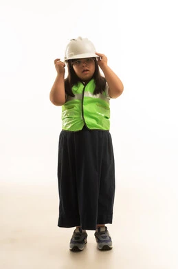 بنت صغيرة عربية خليجية سعودية تعاني من متلازمة داون تنقر على خوذة الامان البيضاء , ارتداي زي مدري موحد لون رمادي و سترة واقية عاكسة للضوء , ايماءات التفكير و التساؤل , النظر الى شئ ما , الجسم بالطول الكامل بورتريه خلفية بيضاء