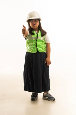 Standing in front of the camera with a thumbs-up sign indicating approval and admiration, a young Arab girl from the Gulf, Saudi, who has Down syndrome, wearing a gray school uniform and an architect's uniform, full-body shot, following safety and protection instructions, portrait with a white background.