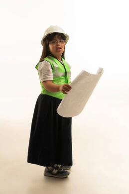 Looking at the camera while holding a large architectural plan, a small Arab Gulf Saudi girl with Down syndrome wearing a reflective safety vest and a white safety helmet, dressed in a gray school uniform, full body, white background.
