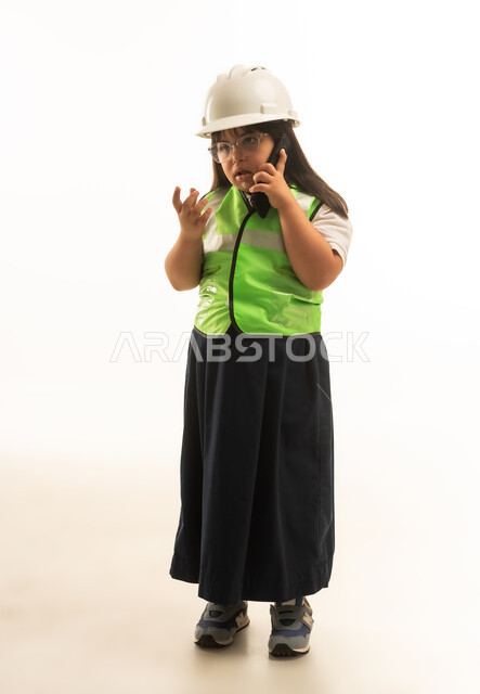 Talking on a mobile phone, expressions of confusion and questioning, a small Arab Gulf Saudi girl with Down syndrome wearing an engineer's jacket and a gray school uniform, full-body, portrait with a white background.