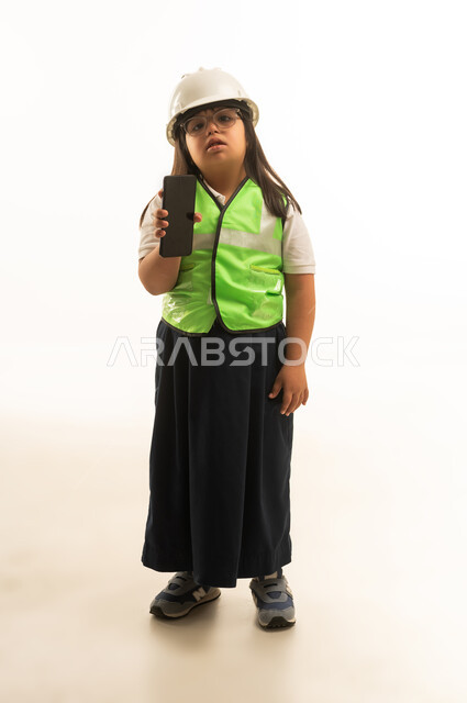 A small Arab Gulf Saudi girl with Down syndrome wearing a gray school uniform with a protective jacket, showing a blank black mobile phone screen to the camera, full body, portrait with a white background.