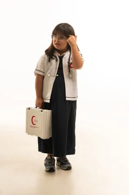 ايماءات التفكير العميق , بنت صغيرة عربية خليجية سعودية من ذوي الهمم ترتدي الزي المدرسي الموحد لون رمادي مع المعطف الطبي الابيض حمل صندوق الاسعافات الاولية , الجسم بالطول الكامل , بورتريه خلفية بيضاء