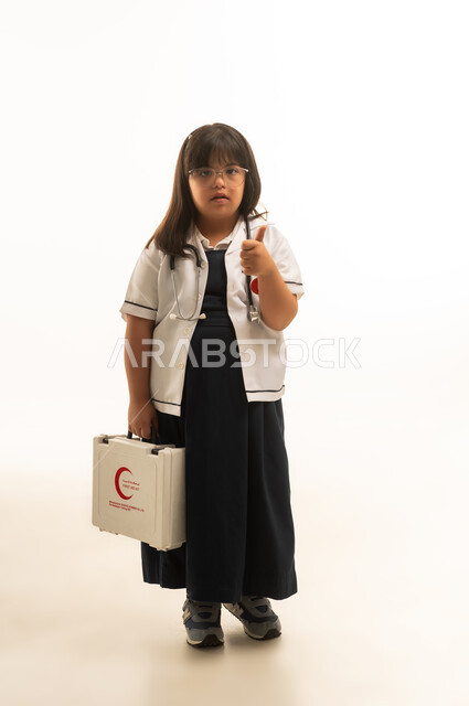 A small Arab Gulf Saudi girl with special needs wearing a white medical coat and a gray school uniform raises her thumb up as a sign of approval and admiration for the medical and educational system, standing in front of the camera, full-body shot, portrait with a white background.