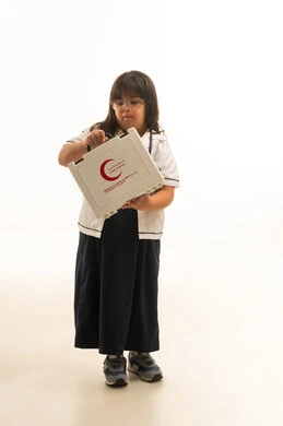 النظر الى صندوق الاسعافات الاولية بين اليدين , بنت صغيرة عربية خليجية سعودية من ذوي الهمم ترتدي معطف طبي ابيض و زي مدرسة موحد لون رمادي تقف امام الكاميرا , الجسم بالطول الكامل , بورتريه خلفية بيضاء