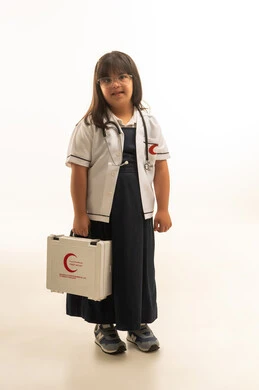 النظر الى الكاميرا بتعابير الابتسامة , بنت صغيرة عربية خليجية سعودية من ذوي الهمم ترتدي زي مدرسة موحد لون رمادي مع المعطف الطبي الابيض , وضع سماعة الطبيب حول الرقبة , الجسم بالطول الكامل , خلفية بيضاء