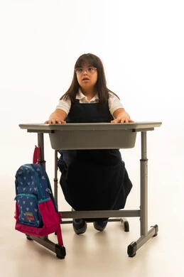 وضع الذراعين على الديسك المدرسي , بنت صغيرة خليجية سعودية تعاني من متلازمة داون ترتدي زي مدرسي موحد لون كحلي تنظر الى شئ ما , بورتريه خلفية بيضاء , الجسم بالطول الكامل