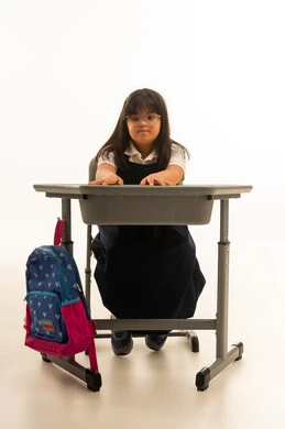 فتح الكفين على الطاولة في المدرسة , بنت صغيرة عربية خليجية سعودية من متلازمة داون ترتدي زي مدرسي موحد لون كحلي تفرد ذراعيها على الديسك , الجسم بالطول الكامل , بورتريه خلفية بيضاء