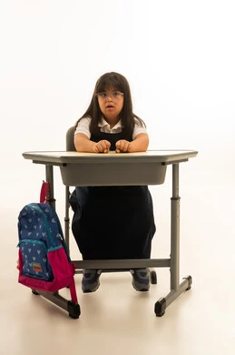 النظر الى الكاميرا مع قفل قبضتي اليد على الطاولة المدرسية , بنت صغيرة عربية خليجية سعودية من متلازمة داون ترتدي زي مدرسي موحد لون كحلي تجلس داخل الفصل , الجسم بالطول الكامل , بورتريه خلفية بيضاء