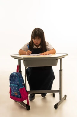 كتابة الواجبات المنزلية و الاداءات الصفية بالقلم الجاف الازرق , طفلة عربية خليجية سعودية صغيرة تعاني من متلازمة داون تجلس على المقعد المدرسي , الجسم بالطول الكامل , بورتريه خلفية بيضاء