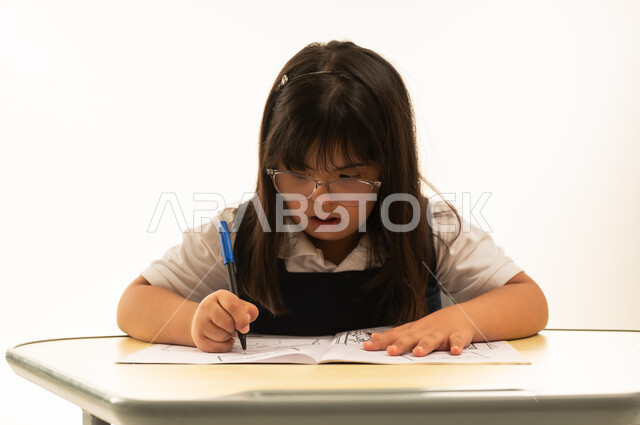 عمل انشطة مسلية اثناء اليوم الدراسي , بنت صغيرة عربية خليجية سعودية من ذوي الهمم ترتدي زي مدرسة موحد لون كحلي تجلس امام طاولة الرسم و التلوين , انشطة ابداعية , بورتريه مقرب , خلفية بيضاء