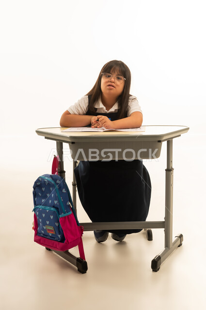 Looking at the camera, a small Arab Gulf Saudi girl with Down syndrome sitting inside the classroom, full-body shot, portrait with a white background, a colorful school bag hanging on the chair.