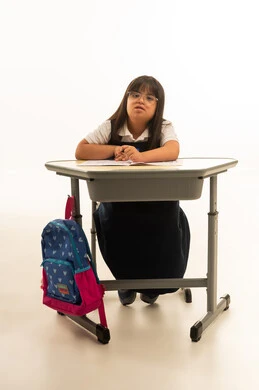 النظر الى الكاميرا , بنت صغيرة عربية خليجية سعودية من متلازمة داون تجلس داخل الفصل الدراسي , الجسم بالطول الكامل , بورتريه خلفية بيضاء , الحقيبة المدرسية الملونة معلقة على المقعد