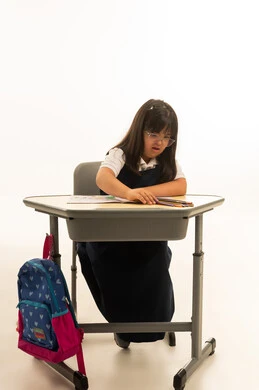 الجلوس داخل الفصل الدراسي , بنت صغيرة عربية خليجية سعودية من متلازمة داون ترتدي زي مدرسة موحد لون كحلي ترسم بالالوان الخشبية , الجسم بالطول الكامل , بورتريه خلفية بيضاء