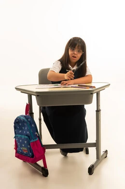 النظر للكاميرا , بورتريه خلفية بيضاء , بنت صغيرة عربية خليجية سعودية من ذوي الهمم ترتدي زي مدرسة موحد لون كحلي تمسك القلم الاسود الخشبي , الجلوس داخل الصف الدراسي , الجسم بالطول الكامل