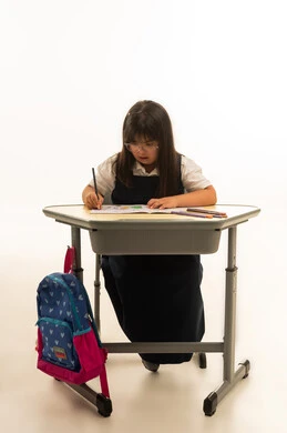 The colorful school bag is hanging on the chair, a small Arab Gulf Saudi girl with Down syndrome is wearing a uniform in navy blue sitting inside the classroom, focusing on writing her homework, portrait with a white background, full body.