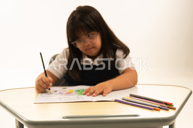 تحسين الوظائف الذهنية لذوي الاحتياجات الخاص باستخدام الرسم و التلوين , بنت صغيرة عربية خليجية سعودية تعاني من متلازمة داون ترتدي زي مدرسة موحد لون كحلي تمسك اللون الخشبي , الجلوس داخل الصف الدراسي , قضاء اوقات ممتعة , بورتريه مقرب , خلفية بيضاء