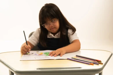 تحسين الوظائف الذهنية لذوي الاحتياجات الخاص باستخدام الرسم و التلوين , بنت صغيرة عربية خليجية سعودية تعاني من متلازمة داون ترتدي زي مدرسة موحد لون كحلي تمسك اللون الخشبي , الجلوس داخل الصف الدراسي , قضاء اوقات ممتعة , بورتريه مقرب , خلفية بيضاء