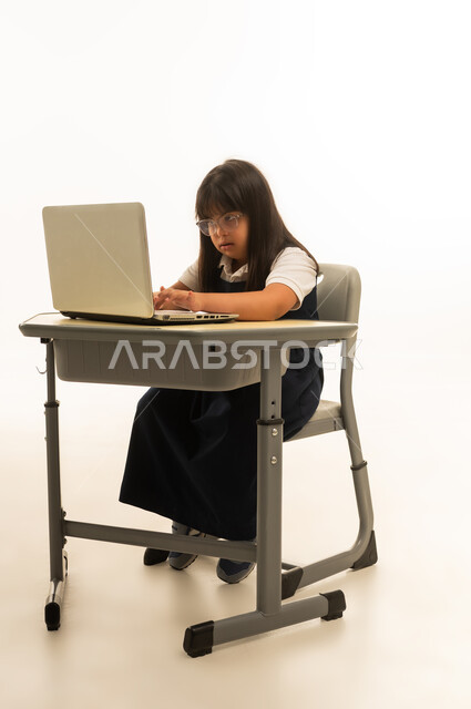 Writing class performances and homework using a mobile computer, a young Arab Gulf Saudi girl with special needs wearing a navy blue school uniform looks intently at the computer screen, utilizing modern technological devices within the Saudi education system, full-body, portrait with a white background.