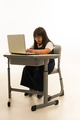 كتابة الاداءات الصفية و الواجب المنزلي باستخدام جهاز الحاسب الالي المتنقل , بنت صغيرة عربية خليجية سعوديو من ذوي الهمم ترتدي الزي المدرسي الموحد لون كحلي تنظر بتركيز الى شاشة الحاسوب , الاستعانة بالاجهزة التقنية الحديثة داخل نظام التعليم السعودي , الجسم بالطول الكامل , بورتريه خلفية بيضاء