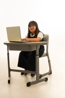 Typing on a laptop keyboard, sitting inside a classroom, a young Arab Gulf Saudi girl with Down syndrome wearing a navy blue school uniform, gestures of happiness and smiling, portrait with a white background, full body, looking at the camera.