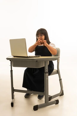 النظر الى شاشة الحاسب الالي المتنقل , تشكيل علامة القلب باليدين و رفعها بالهواء , بنت صغيرة عربية خليجية سعودية من ذوي الهمم ترتدي الزي المدرسي الموحد لون كحلي , الجلوس داخل الصف الدراسي , الجسم بالطول الكامل , بورتريه خلفية بيضاء