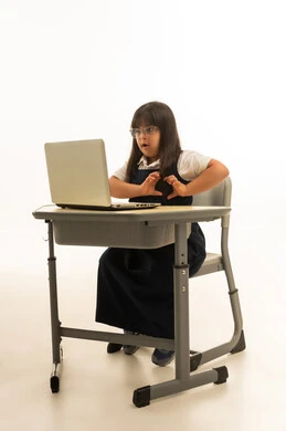 Heart sign with hands, a small Arab Gulf Saudi girl with special needs wearing a uniform school outfit in navy blue sitting inside a classroom, using a laptop in the educational system, looking at the computer, full body, portrait with a white background.