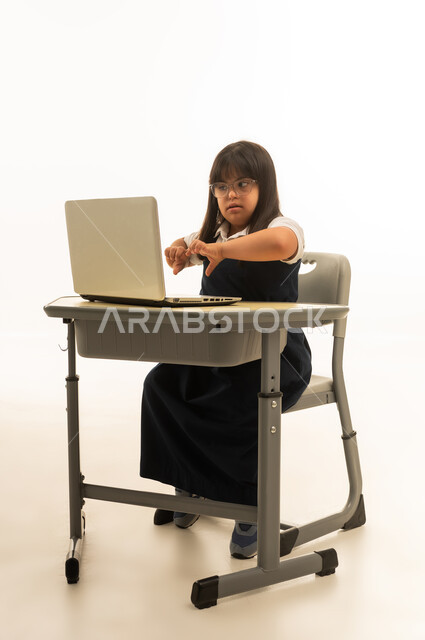 Joining hands in front of a mobile computer with a heart sign, a small Arab Gulf Saudi girl with special needs wearing a uniform teacher's outfit in navy blue, using modern devices in the Saudi education system, full-body, portrait with a white background.