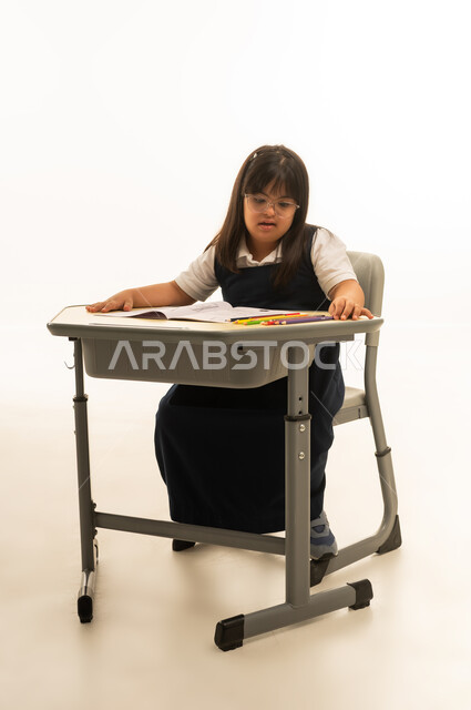 النظر الى الالوان الخشبية على مقعد المدرسة لاختيار اللون المناسب , بنت صغيرة عربية خليجية سعودية تعاني من متلازمة داون ترتد زي المدرسة الموحد لون الكحلي , الجلوس داخل الصف المدرسي , الجسم بالطول الكامل , بورتريه خلفية بيضاء