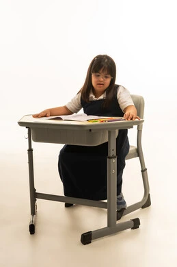 Looking at the wooden colors on the school bench to choose the right color, a small Arab Gulf Saudi girl with Down syndrome wearing the school uniform in navy blue, sitting inside the classroom, full-body, portrait with a white background.