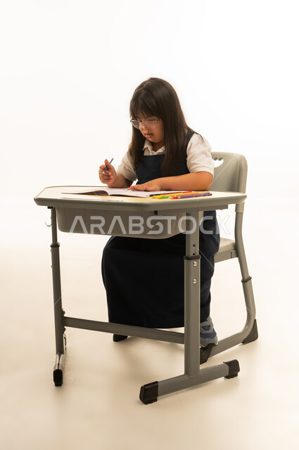 انشطة ابداعية تساعد ذوي الاحتياجات الخاصة على تنمية المهارات الذهنية , بنت صغيرة عربية خليجية سعودية من متلازمة داون ترتد زي المدرسة الموحد لون كحلي تجلس داخل الصف المدرسي , النظر بتركيز الى الكراسة , الجسم بالطول الكامل , بورتريه خلفية بيضاء