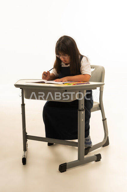 النظر بتركيز شديد الى الكراسة على المقعد المدرسي , بنت صغيرة عربية خليجية سعودية من متلازمة داون ترتدي زي المدرسة الموحد تجلس على مقعد الصف , بورتريه الجسم بالطول الكامل , خلفية بيضاء