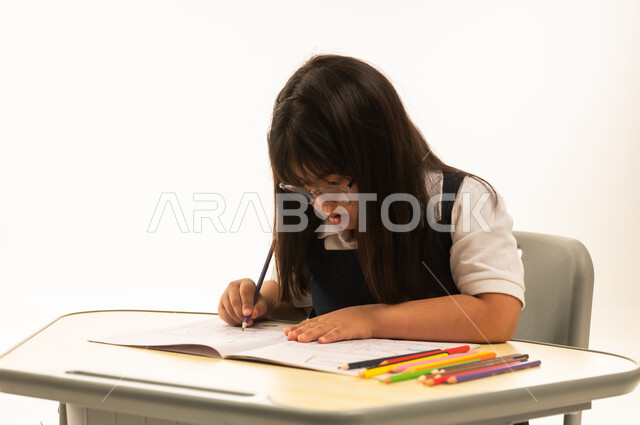 الرسم و التلوين باستخدام الالوان الخشبية , بنت صغيرة عربية خليجية سعودية من ذوي الهمم ترتدي زي المدرسة الموحد لون كحلي تجلس داخل الصف المدرسي , بورتريه مقرب , خلفة بيضاء , الاعتماد على الانشطة الابداعية بالمنظومة التعليمية