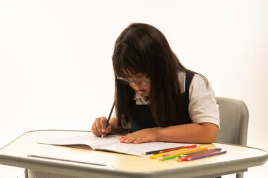 الرسم و التلوين باستخدام الالوان الخشبية , بنت صغيرة عربية خليجية سعودية من ذوي الهمم ترتدي زي المدرسة الموحد لون كحلي تجلس داخل الصف المدرسي , بورتريه مقرب , خلفة بيضاء , الاعتماد على الانشطة الابداعية بالمنظومة التعليمية