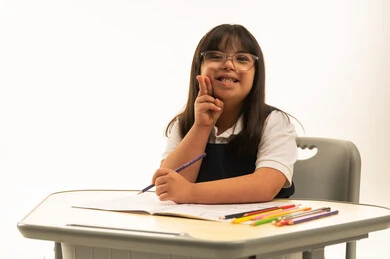 Two fingers on the cheek and a smile in front of the camera, a small Arab Gulf Saudi girl with Down syndrome wearing a navy blue school uniform, sitting on school benches, using a pencil and colored pencils, having fun, close-up portrait, white background.