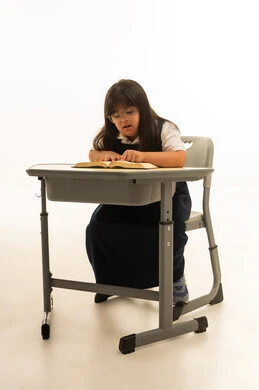 A small Arab Gulf Saudi girl with Down syndrome sitting in a classroom, inclusion of people with disabilities in the educational system, looking at an open book, full-length body, portrait with a white background.