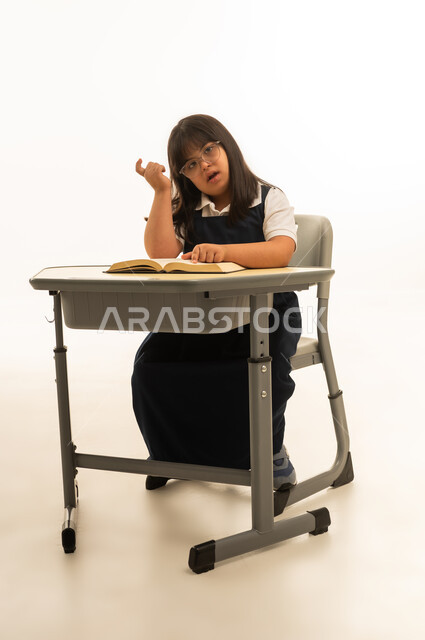 Looking at the camera with a raised index finger as a sign of thinking, a small Arab girl from the Gulf, Saudi, with special needs, wearing a school uniform, sitting inside the classroom, portrait with a white background, full body.