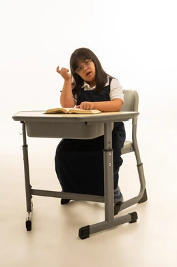 Looking at the camera with a raised index finger as a sign of thinking, a small Arab girl from the Gulf, Saudi, with special needs, wearing a school uniform, sitting inside the classroom, portrait with a white background, full body.