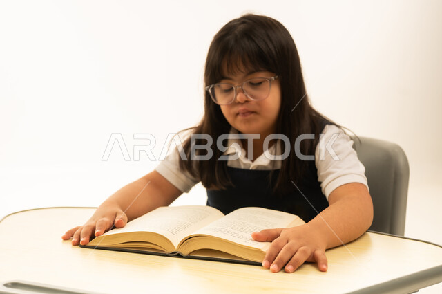 قراءة الكتاب المفتوح اثناء اليوم الدراسي , بنت صغيرة عربية خليجية سعودية من متلازمة داون ترتدي زي المدرسة الموحد , بورتريه مقرب , خلفية بيضاء