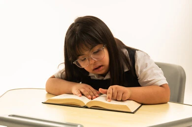 لمس الكتاب المدرسي بالاصابع , بنت صغيرة عربية خليجية سعودية تعاني من متلازمة داون ترتدي زي المدرسة الكحلي الموحد , الجلوس داخل الفصل الدراسي , بورتريه مقرب , خلفية بيضاء