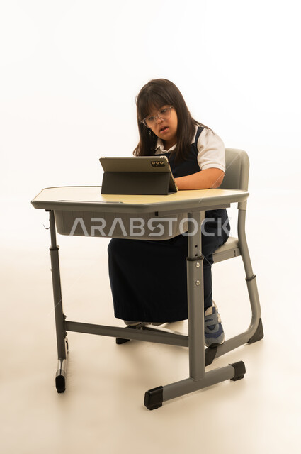 Daily lessons follow-up on the tablet, a small Arab Gulf Saudi girl with determination wearing a navy school uniform sitting inside the classroom, full-body, portrait with a white background.