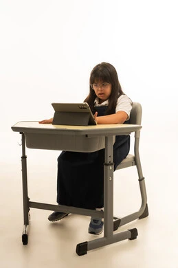 الاعتماد على الاجهزة اللوحية في المنظومة التعليمية , بنت صغيرة عربية خليجية سعودية من ذوي الهمم ترتدي الزي المدرسي الكحلي تجلس داخل الفصل الدراسي , الجسم بالطول الكامل , النظر الي شاشة الجهاز التقني , بورتريه خلفية بيضاء