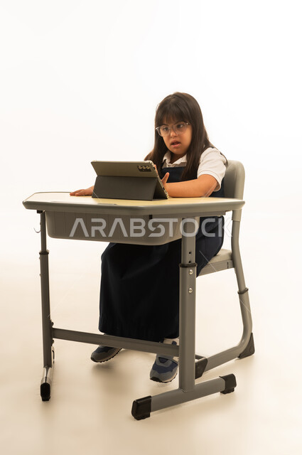 النظر الى شاشة الجهاز اللوحي , بنت عربية خليجية سعودية صغيرة تعاني من متلازمة داون ترتد الزي المدرسي تجلس داخل الفصل الدراسي , استخدام الاجهزة التقنية الحديثة داخل المدارس , الجسم بالطول الكامل , بورتريه خلفية بيضاء