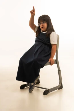 رفع اصبع السبابة لاعلى اشارة الى الطلب و الاستئذان , بنت صغيرة عربية خليجية سعودية من ذوي الهمم ترتدي زي المدرسة الكحلي تجلس على مقعد الفصل , الجسم بالطول الكامل , بورتريه خلفية بيضاء