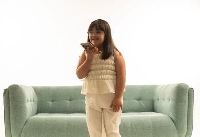 Expressions of happiness and joy while talking on a loudspeaker, a small Arab Gulf Saudi girl with determination holding a mobile phone, looking at something, standing in front of a gray sofa, close-up portrait with a white background.