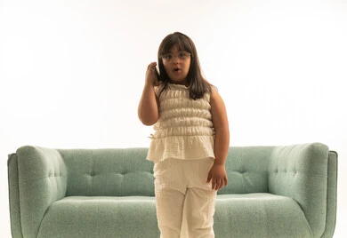 التحدث في الهاتف المحمول , بنت صغيرة عربية خليجية سعودية تعاني من متلازمة داون ترتدي الزي الكاجوال تقف امام اريكة رمادية اللون , بورتريه خلفي بيضاء , النظر للكاميرا