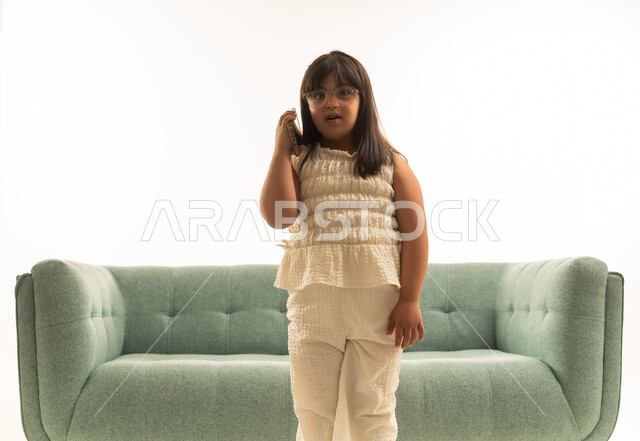 A small Arab Gulf Saudi girl with special needs wearing beige casual attire, standing in front of a gray sofa, listening to a phone call, portrait with a white background, looking at the camera.