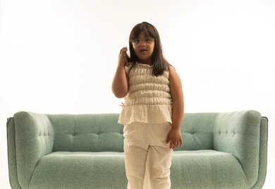 بنت صغيرة عربية خليجية سعودية من ذوي الهمم ترتدي الزي الكاجوال البيج , الوقوف امام اريكة رمادية اللون  , الاستماع الى المكالمة الهاتفية , بورتريه خلفية بيضاء , النظر للكاميرا