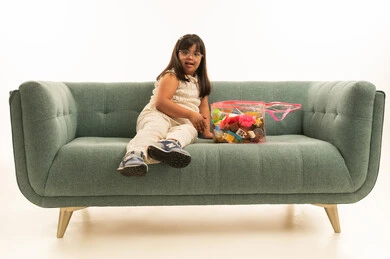 ايماءات الابتسامة و النظر للكاميرا , بنت صغيرة عربية خليجية سعودية من ذوي الهمم ترتدي الزي الكاجوال تجلس على اريكة رمادية اللون . صندوق العاب بلاستيكي شفاف , الجسم بالطول الكامل , بورتريه خلفية بيضاء