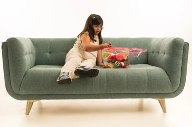 تصفيط الالعاب المبعثرة على اريكة رمادية اللون , بنت صغيرة عربية خليجية سعودية تعاني من متلازمة داون ترتدي الزي الكاجوال , بورتريه خلفية بيضاء , الجسم بالطول الكامل